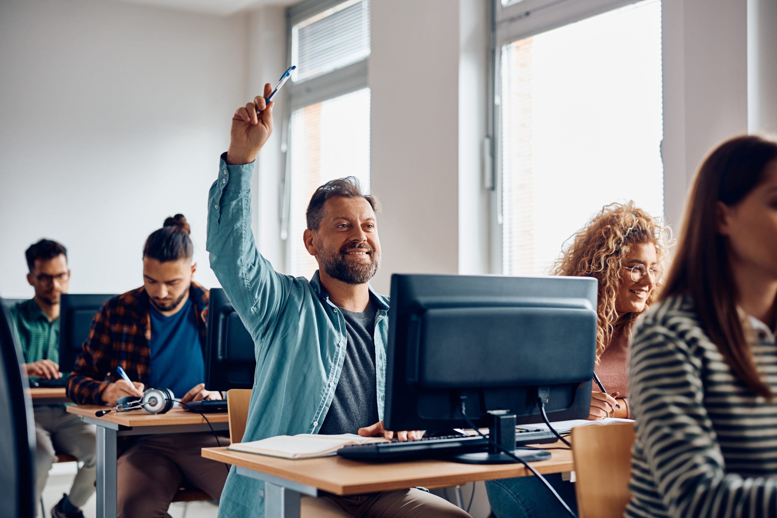A smiling man raising his hand to answer a question in a computer class.
