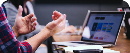 Hands of a person wearing a red and blue plaid shirt. Person is sitting at a desk in front of a laptop with a book open and appear to be gesticulating with their hands as if talking.
